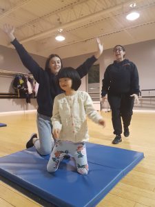 Teacher supervising little girl and a female volunteer who are dancing on a mat.