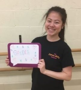 Sandra smiles and holds a whiteboard with her name. 