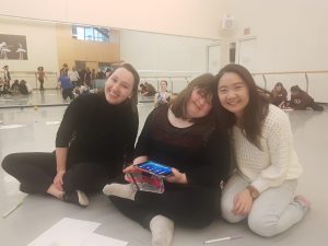 Three women sitting on a dance studio floor and smilling. The one in the middle is holding and iPad.